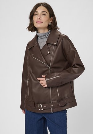 Frau in einer dunkelbraunen, oversized Ledermantel mit silbernen Reißverschlüssen und Gürtel, grauem Rollkragenpullover und blauen Jeans vor einem schlichten Hintergrund.