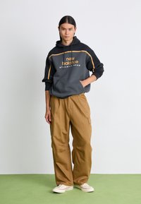 Gray hoodie with yellow accents and "new balance" logo, paired with khaki cargo pants, and white sneakers on green floor background.