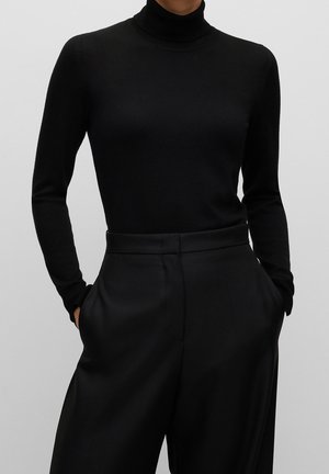 Personne portant un pull à col roulé noir à manches longues, rentré dans un pantalon noir taille haute à jambes larges, les mains dans les poches.