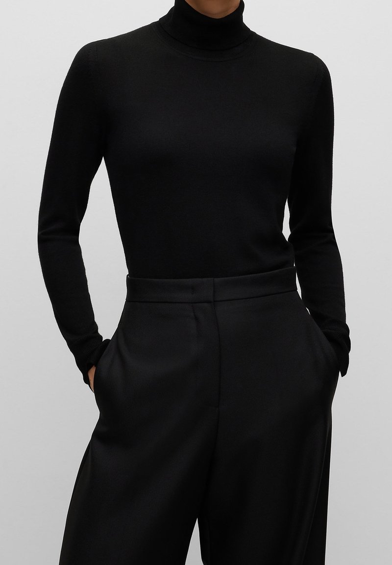 Personne portant un pull à col roulé noir à manches longues, rentré dans un pantalon noir taille haute à jambes larges, les mains dans les poches.