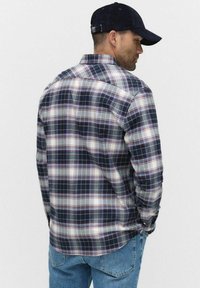 Blauw-wit geblokt flanellen shirt met rode accenten; lange mouwen, met knoopkraag; gedragen met denim jeans en een donkere pet.