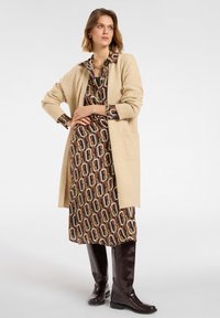 Cardigan a maglia color beige con maniche lunghe sopra un vestito marrone a fantasia con forme collegate, abbinato a stivali lucidi marroni al ginocchio.