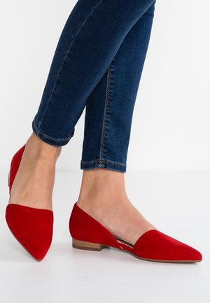 Pieds chaussés de ballerines pointues rouge vif associées à un jean skinny bleu foncé sur un fond blanc uni.