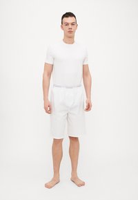 Weißes Kurzarmhemd und passende Shorts, beide aus glattem Stoff. Die Shorts haben einen elastischen Bund mit einem Logo. Modell barfuß.