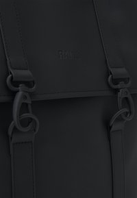 Sac à dos noir imperméable avec sangles réglables, doté de fermetures à pression, d'une texture lisse et du logo "RAINS" en relief à l'avant.