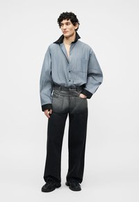 Νεαρός άνδρας με υπερμεγέθη μπλε ριγέ πουκάμισο με μαύρο γιακά και μανσέτες, συνδυασμένο με δίχρωμο μαύρο και γκρι παντελόνι φαρδιάς γραμμής και μαύρα παπούτσια.