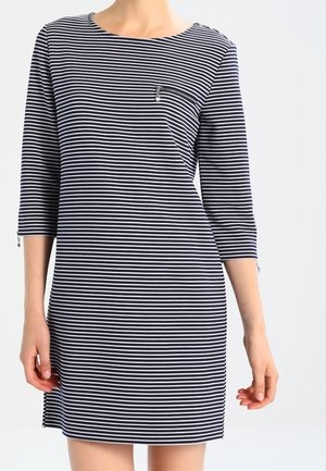 Femme portant une robe à rayures horizontales bleu marine et blanche, avec des manches trois-quarts et une petite poche zippée sur la poitrine.