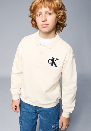 Kermanvärinen neule, jossa on kaulus ja vasemmassa rinnassa musta brodeerattu "cK" -logo, yhdistettynä sinisiin cargo-farkkuihin.