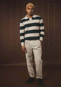 Pull en maille rayée vert foncé et crème, pantalon beige, et chaussures noires. L'arrière-plan présente un éclairage doux avec des guirlandes lumineuses.