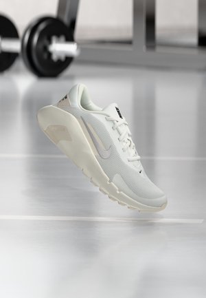 Chaussure de course Nike blanche flotte au-dessus du sol de la salle de sport avec le matériel de musculation flou en arrière-plan.