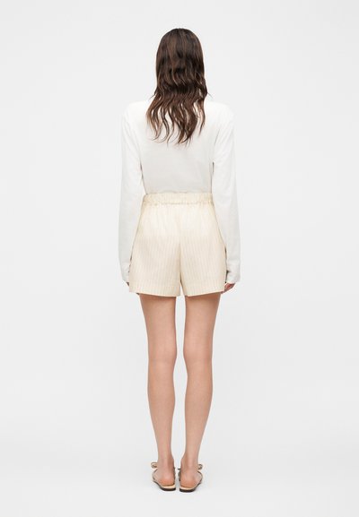 Γυναίκα με μακριά κυματιστά μαλλιά που φοράει λευκή μπλούζα με μακριά μανίκια, μπεζ κοντό σορτς με ρίγες στη μέση και μπεζ ανοιχτά σανδάλια με φλατ σόλα, που στέκεται με την πλάτη προς τα εμπρός.