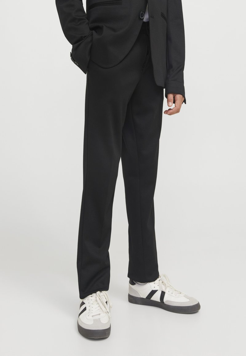 Personne portant un pantalon de costume noir et des baskets blanches avec des bandes noires, debout devant un fond uni.