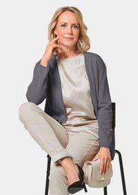 Grijze cardigan over een licht satijnen top, gecombineerd met beige broek. Zwarte hoge hakken en een kleine crèmekleurige handtas met gouden hardware. Zittende houding.