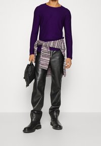 Lila långärmad skjorta, svarta läderbyxor och svarta stövlar. Ett randigt stickat plagg knutet runt midjan; en väska i handen.