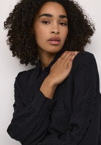 Bluză neagră cu detalii delicate din dantelă pe mâneci și pe plăcuța frontală. Are un guler clasic și închidere cu nasturi. Material moale, texturat.