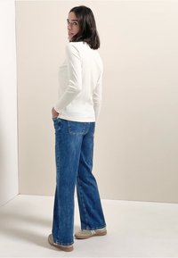 Haut blanc à manches longues côtelé associé à un jean bleu coupe décontractée. Le jean comporte des poches arrière et un léger évasement à l'ourlet. Chaussures décontractées visibles.