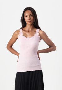Débardeur rose en maille côtelée avec un décolleté en dentelle festonnée et des bretelles, design ajusté, tissu léger, associé à une jupe plissée noire.