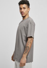 Szürke, oversized póló, puha anyagból, kerek nyakkivágással és rövid ujjakkal, zöld címkével az alján és tetoválással a karon.