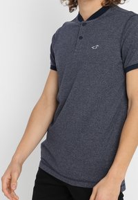 Marinblå polo skjorta med korta ärmar, texturerad tyg och två-knappsstängning. Har en liten logo på bröstet och kontrasterande krage.