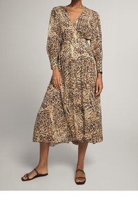 Długa sukienka kopertowa w beżowo-brązowy wzór zwierzęcy, z długimi rękawami i wcięciem w talii, z lejącą spódnicą i płaskimi czarnymi sandałami.