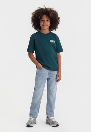 Bambino con capelli ricci che indossa una maglietta New Balance verde scuro, jeans azzurri, scarpe da ginnastica grigie, in piedi con una mano in tasca.