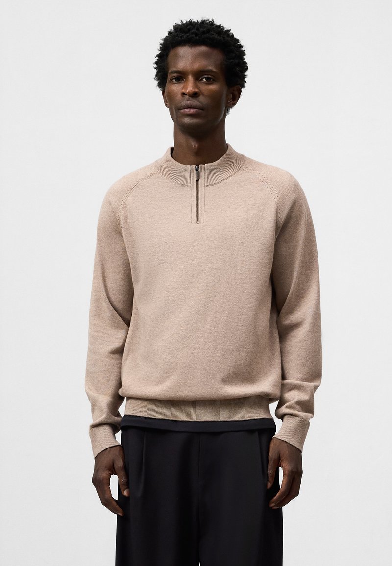 Maglione beige a maglia con colletto a mezza zip e polsini a costine. Presenta una vestibilità rilassata e una texture fine, abbinato a pantaloni scuri.