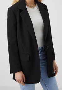 Musta säädöissä suunniteltu bleiseri, jossa on yksinkertainen nappilukko, notched kaulukset ja sileä pinta, yhdistetty valkoiseen ribbipaita.