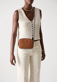 Braune strukturiert Leder-Crossbody-Tasche mit silberner Kettenriemen. Verfügt über ein dreieckiges Logo auf der Vorderseite, kombiniert mit einem cremefarbenen Strickoberteil und hellen Hosen.