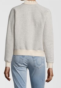 Sudadera gris claro con cuello y puños acanalados, que presenta una superficie ligeramente texturizada. Combinada con jeans de un azul medio que muestran detalles de costura.