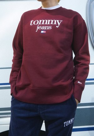 Sudadera burdeos con texto y logo blancos de "tommy jeans", con cuello redondo, puños y dobladillo acanalados. Combinada con pantalones azul marino.