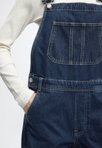 Tumesinise pesuga teksariidest üleõlakatted, millel on reguleeritavad metallist rihmad, eesmine tasku ja küljetaskud, sileda viimistluse ja lõdva lõikega.