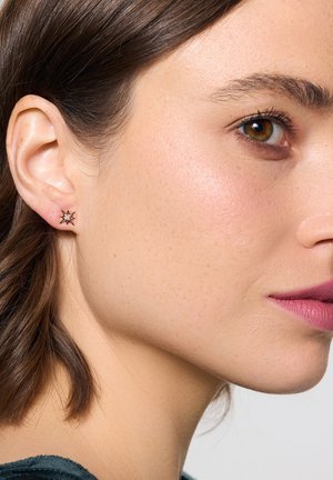 Złote kolczyki w kształcie gwiazdy z przezroczystym kamieniem w centrum, noszone w lewym uchu, prezentujące gładką fakturę i błyszczące wykończenie.