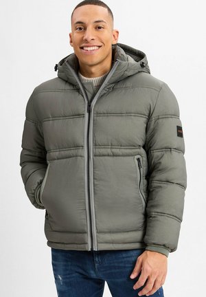 Lächelnder Mann in einer grauen gepolsterten Jacke mit Kapuze, Frontreißverschluss und seitlichen Reißverschlusstaschen, kombiniert mit blauen Jeans.