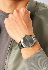 Silberner Edelstahl-Armbanduhr mit grauem Zifferblatt, strukturiertem Gehäuse und Gliederarmband, getragen an einem Handgelenk, dessen hellgrauer Jackenärmel sichtbar ist.