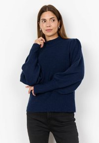 Pull en maille côtelée marine avec un col montant, des manches bouffantes et une coupe décontractée. Le tissu a une texture douce ; légèrement court au niveau de la taille.