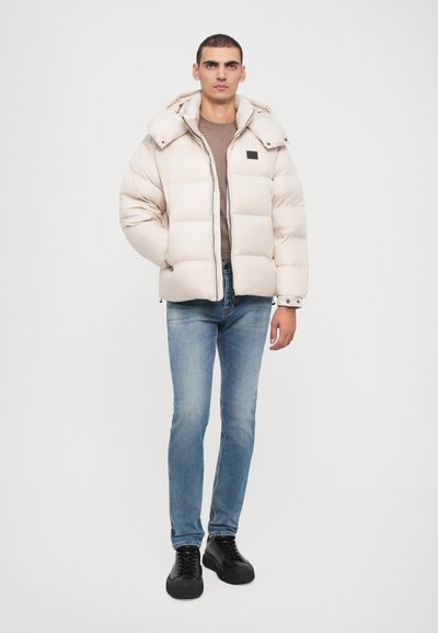 Pufferjakke i lys beige med hætte, med quiltet design og et sort logo-flag. Båret over en brun top og blå jeans.