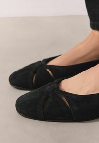 Fekete velúr balettcipő lekerekített orral, keresztpántos kialakítással és kivágott részletekkel. Texturált felület és alacsony profilú talp.