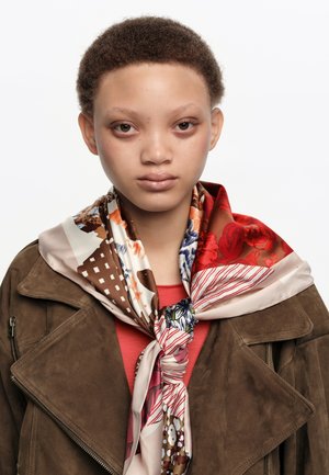 Persona joven con cabello corto y rizado que lleva una chaqueta de ante marrón y una bufanda colorida con estampado atada al cuello.