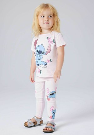 Young blonde girl wearing pink Stitch-themed pajamas and silver sandals, standing against a light gray background.