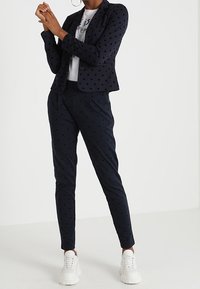 Marinblå polka prickig kavaj och matchande byxor i texturerad tyg, med en figursydd design. Kombinationen med vita sneakers ger en kontrast.