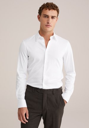 Chemise blanche à manches longues avec col pointu, léger brillant, et boutons sur le devant, associée à un pantalon gris foncé taillé sur mesure.