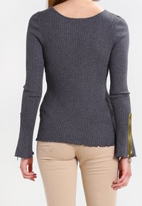 Grå ribbad långärmad topp med rund halsringning och utsvängda ärmar; har kontrasterande dragkedjor vid ärmarna och en rå fåll.