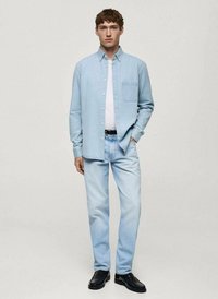 Camicia blu chiaro con bottoni e taschino sul petto, T-shirt bianca sotto, jeans blu chiaro e scarpe nere. Tessuto liscio, vestibilità casual.