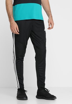 Mężczyzna w czarnych spodniach dresowych Adidas z białymi paskami i czarnych butach sportowych, stojący na jednolitym jasnym tle.