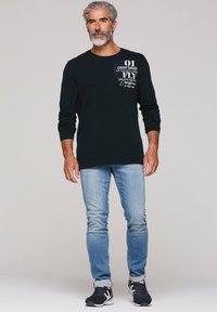Zwart longsleeve t-shirt met een grafische print in wit en blauw, gecombineerd met lichtblauwe jeans en zwarte sportschoenen.