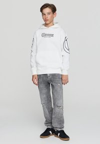 Biała bluza z kapturem i kieszenią z przodu, czarny nadruk tekstowy i wzory na rękawach, w połączeniu z postarzanymi szarymi dżinsami i czarnymi sneakersami.