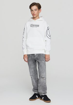 Felpa bianca con cappuccio e tasca frontale, con testo grafico nero e design sulle maniche, abbinata a jeans grigi strappati e sneakers nere.