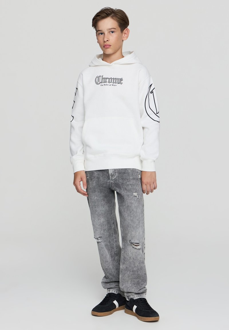 Biała bluza z kapturem i kieszenią z przodu, czarny nadruk tekstowy i wzory na rękawach, w połączeniu z postarzanymi szarymi dżinsami i czarnymi sneakersami.