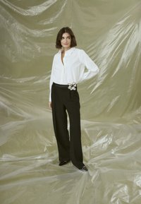 White button-up shirt with a shiny finish and black wide-leg trousers featuring a floral brooch. Background is a wrinkled beige plastic sheet.