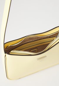 Gul kunstlærveske med en glatt overflate, med flere glidelåslommer, brune detaljer og et trekantet logo.